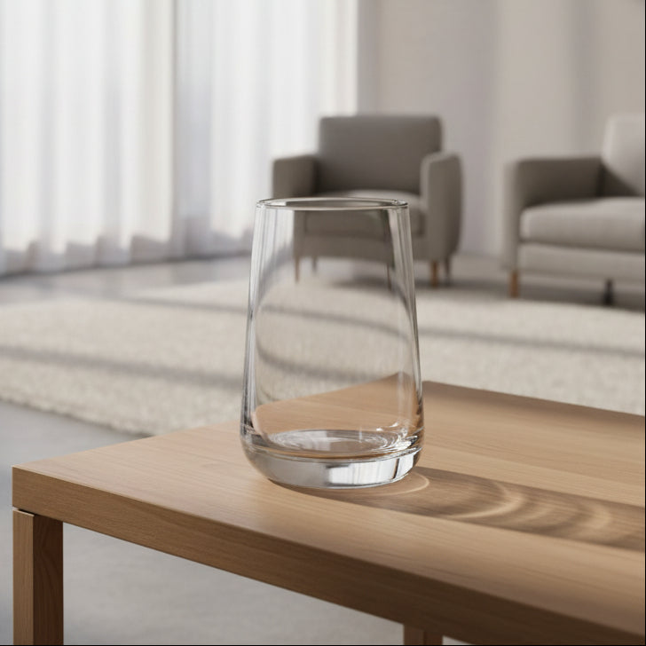 A clear, tapered lowball glass vase with a thick, rounded base, shown on a reflective surface against a light grey background.